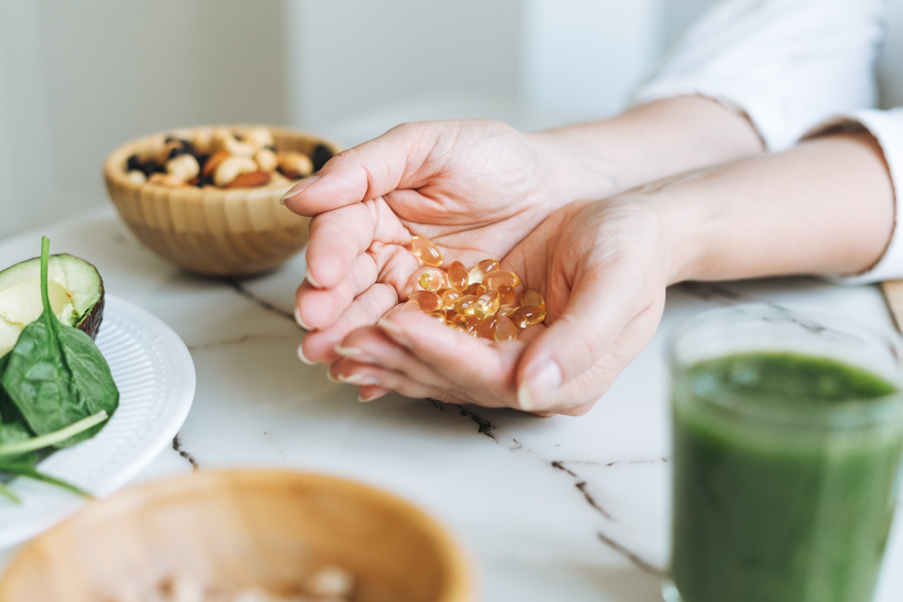 Manos con cápsulas de omega 3 y vitamina D junto a un batido verde y frutos secos