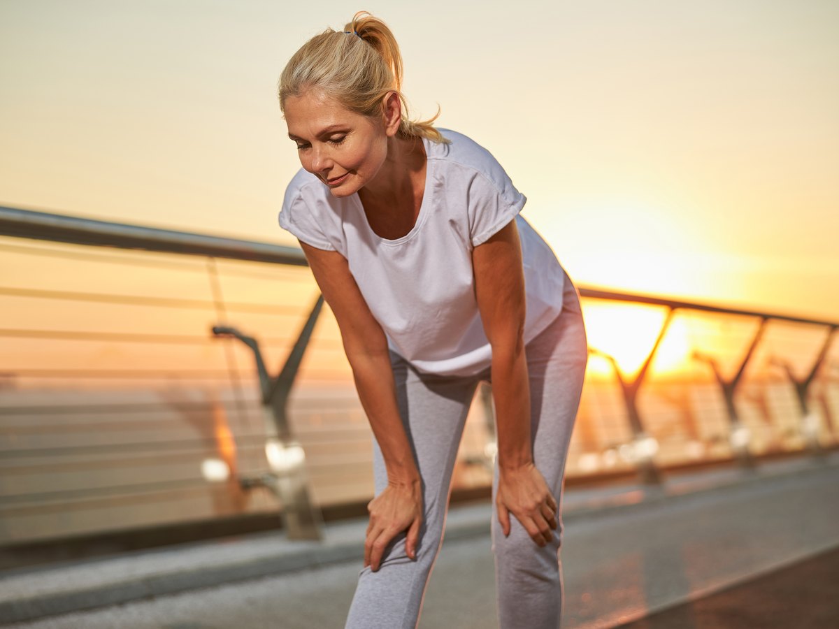 Mujer de mediana edad recuperando el aliento con las manos en las rodillas tras hacer ejercicio al aire libre al amanecer